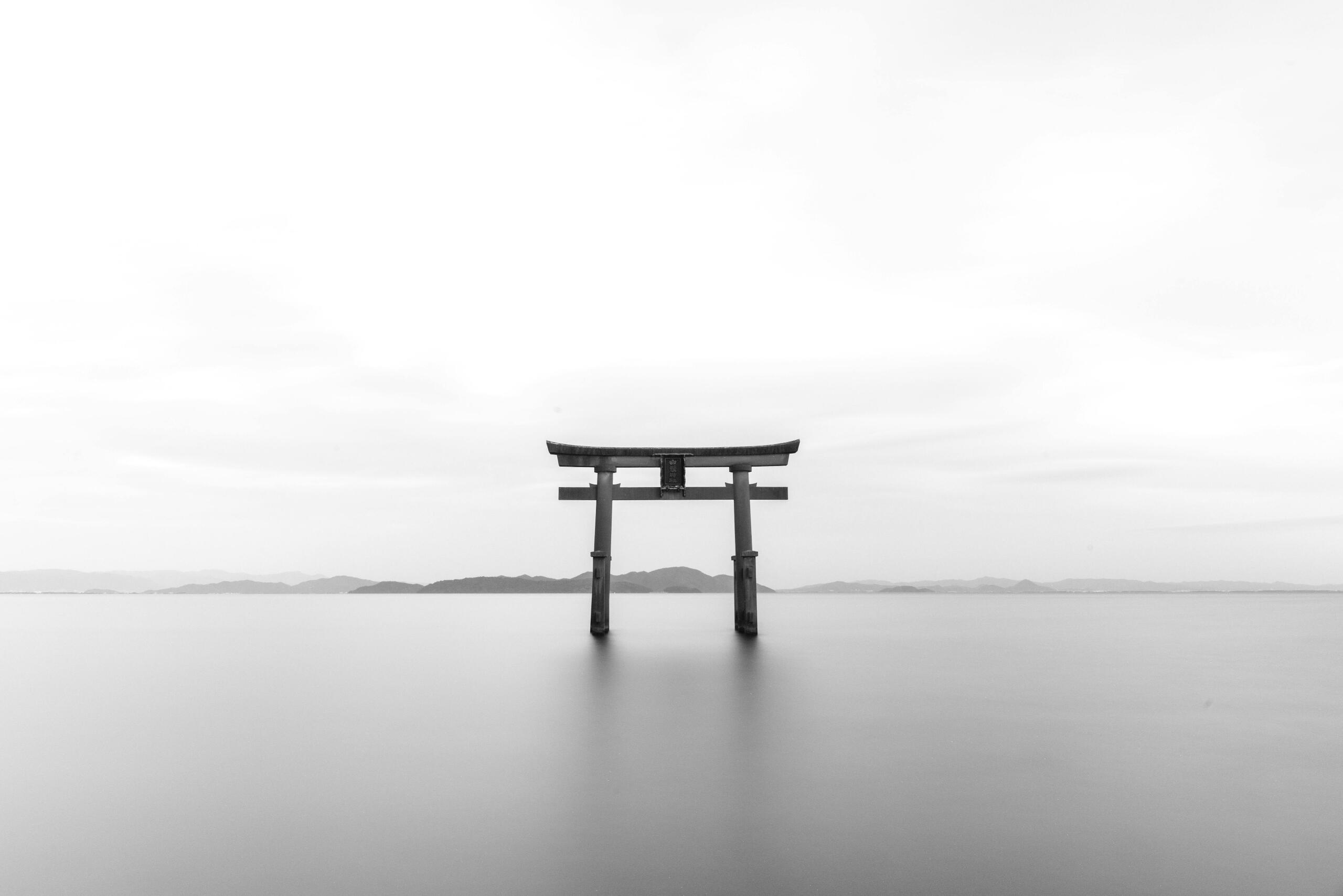 A black & white photo of a torii gate emerging from the ocean with mountains far in the background.